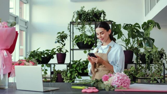 Young, successful female florist receiving an order on her mobile phone. She smiles and selects a few pink carnations and begins to arrange a bouquet.