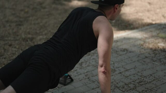 A determined man wearing black performs pushups in a park, using handles for added intensity in his fitness routine. He is working hard for his health and wellbeing.
