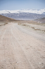 Dusty road of the Pamir Highway winds and twists in the valley of the Tien Shan Mountains against the backdrop of a mountain range with snow in Tajikistan in the Pamirs, landscape for the background
