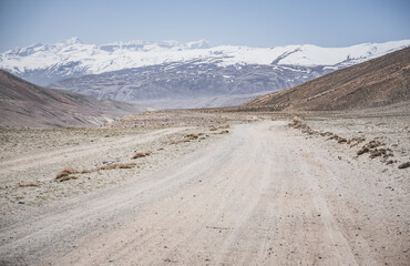 Dusty road of the Pamir Highway winds and twists in the valley of the Tien Shan Mountains against the backdrop of a mountain range with snow in Tajikistan in the Pamirs, landscape for the background