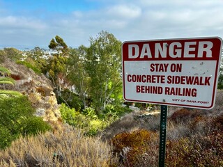 Red warning sign at Dana Point, California, with lush greenery and a cloudy sky in the background, emphasizing safety precautions.