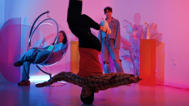 Happy hipster doing head spin while friend looking at performance with neon light dancing studio. Professional caucasian man moving to music while smart people sitting and pose at camera. Regalement.