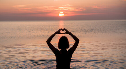 Fototapeta premium A young woman at the beach making a heart shape sign using her hands against the backdrop of a sun