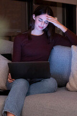 Stressed woman working late at night on a laptop, touching her forehead due to a headache, sitting alone on a sofa in a dark living room, struggling to meet an important work project deadline