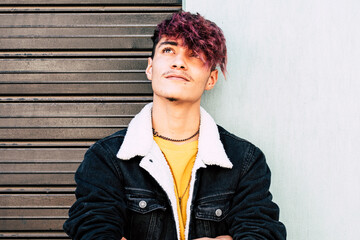 Casual teenage student with colorful hair, wearing denim, stands outside leaning against a wall,...