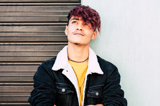 Casual teenage student with colorful hair, wearing denim, stands outside leaning against a wall, looking up thoughtfully. His relaxed and confident demeanor reflects his alternative style and youthful