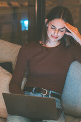 A woman sitting alone on a couch at night, illuminated by the soft glow of her laptop, looking...