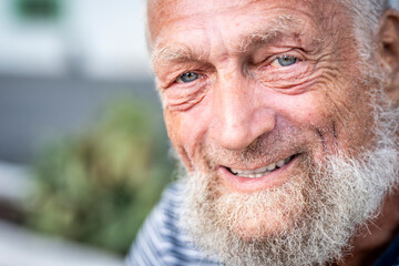 Portrait of an elderly man with blue eyes smiling sadly alone in a park. His wrinkled face shows signs of aging, representing the concept of Alzheimer’s and mature age, where a small, false happiness