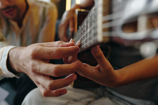 Close-up of hands fretting on guitar strings while another person guides playing technique. No visible faces enhancing learning experience and focusing on guitar frets