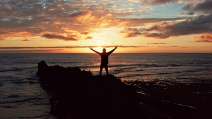 Man stands on a rock against the ocean during summer sunset. Human with raised hands looks to sun over horizon in morning while sunrise. Man feels happy and freedom. Silhouette of a man against  sky