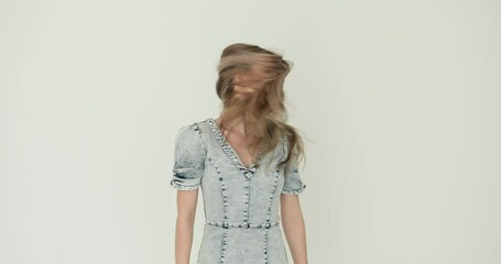 Happy young woman in denim dress shakes head tossing long loose hair side to side on white background. Attractive lady demonstrates stylish hairstyle