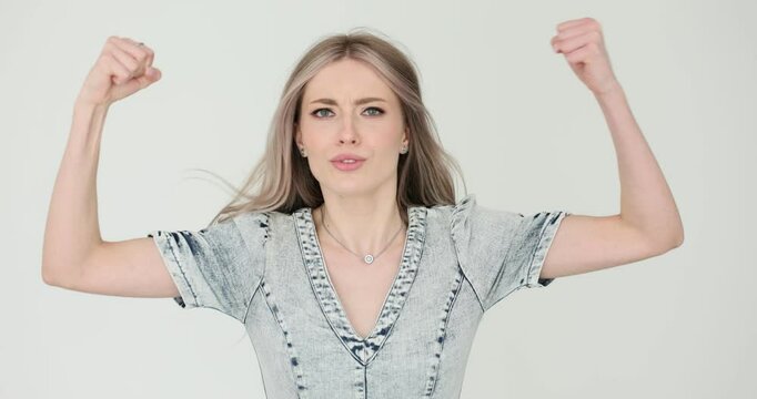 Angry young woman demonstrates irritation waving fists on white background. Indignant blonde female in denim dress with aggressive behaviour