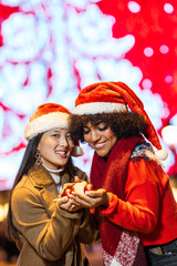 Two friends holding christmas ornaments in front of christmas tree lights