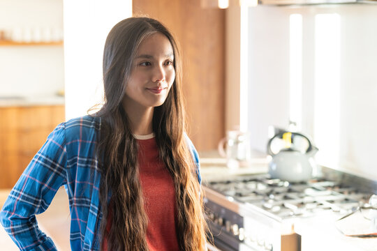 At home, Smiling young asian woman standing in kitchen near stove, wearing casual plaid shirt - Powered by Adobe