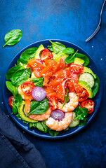 Healthy food salmon salad for keto diet with shrimp, avocado, spinach, cucumber, tomato, cashew, sesame seeds. Blue background, top view