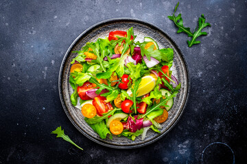 Vegan vegetable salad with tomatoes, cucumber, red onion, lettuce and arugula. Black background, top view
