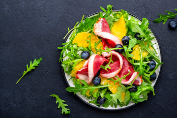 Foodie food salad with raw smoked duck and juicy orange, blueberry and arugula on black background, top view