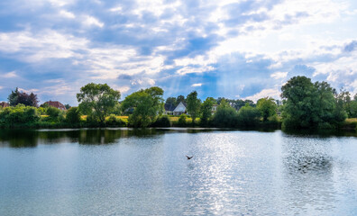 Peaceful lakeside view with trees, houses, and soft sunlight breaking through the clouds