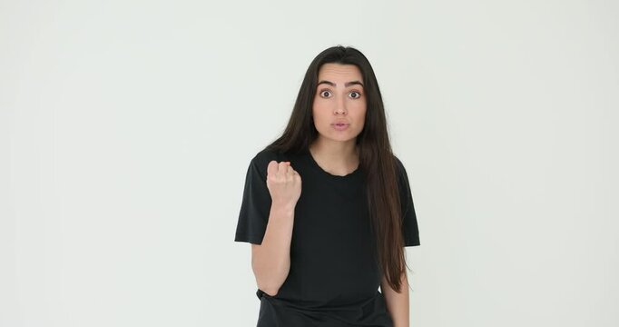 Angry young woman threatens by fist standing on white background. Female model expresses aggression with gesture ready to fight enemy