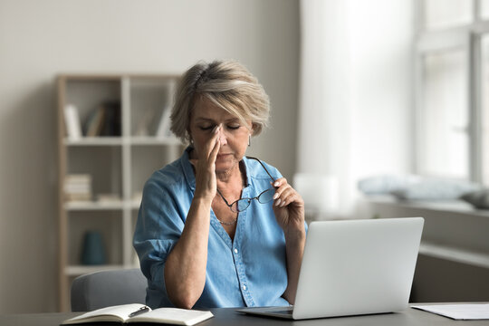 Mature woman takes off eyeglasses sit at desk with laptop, experiences discomfort or tiredness, due to eyestrain, remove glasses, rubbing nose bridge. Digital eye strain, bad influence of modern tech