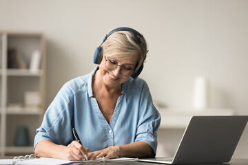 Smiling mature older woman sit at desk working on-line, wear wireless headphones e-learning using...