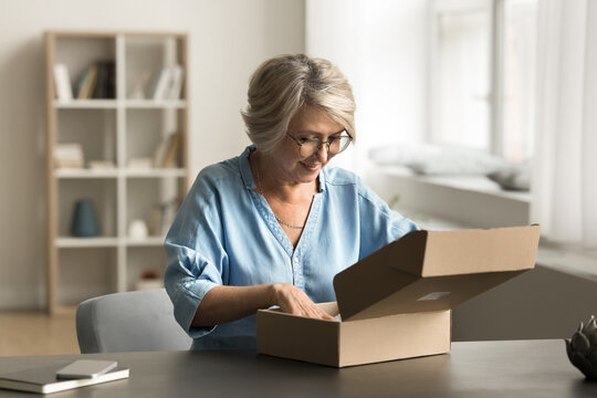 Smiling mature woman sit at table at home opens received parcel, review bought goods for home interior or fashion items enjoy easy, express courier services, ordered trustworthy transportation company