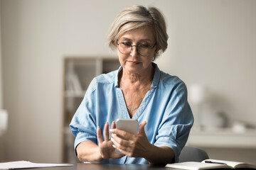 Attractive older woman wear glasses sit at table with smartphone, sending text message, checking notifications, browsing wireless internet, staying connected using modern technology for communication