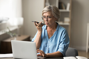 Mature woman using smartphone sit at desk with laptop, engaged in voice search or dictation on...