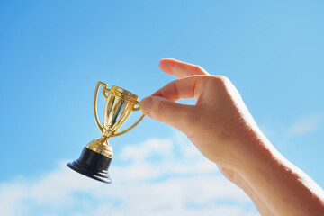 Hand holds and raised small cup trophy, holding cup against clean sky backgrund. Consolation prize...