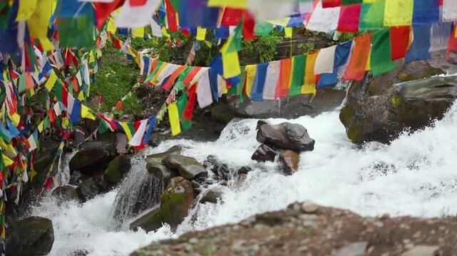4K shot of rapidly flowing river cascade with colorful prayer flags at Lahaul, Himachal Pradesh, India. Clear stream running through stone and splashing water. 