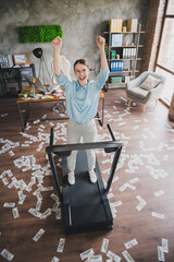 Vertical photo of cheerful lucky lady recruiter dressed shirt flying dollars rising fists running treadmill indoors workplace workstation loft