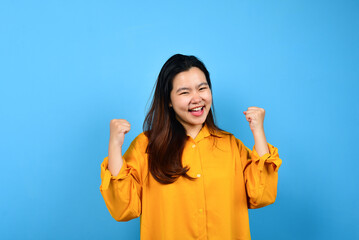 A young Asian woman with a happy successful expression wearing yellow shirt with copy space isolated on blue backround