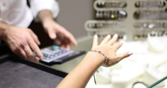 Woman tries on bracelet gems while salesman counts costs on calculator in store slow motion. Female customer buys expensive accessory in jewelry