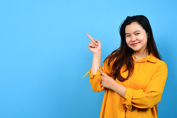 Happy asian woman in orange shirt pointing right side, left side and up side to copy space and looking at the camera isolated blue background