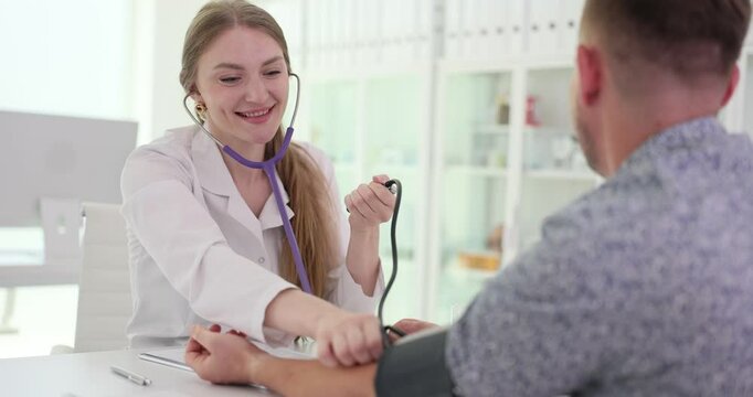 Smiling female doctor measures blood pressure of male patient in clinic. Woman therapist examines man with tonometer at appointment slow motion