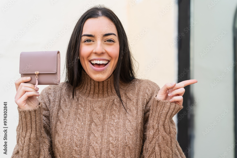 Poster young woman holding a wallet at outdoors surprised and pointing finger to the side