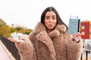 Young woman holding a passport at outdoors making doubts gesture while lifting the shoulders