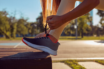 Sporty young woman tying running sneakers in urban park during summertime sunny day.