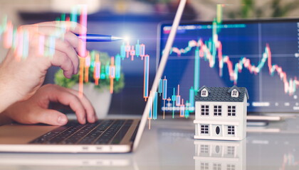 Hands of real estate agent working with laptop at table with house model and candlestick chart