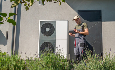 A technician in work overalls checking a heat pump unit with a tablet outside a house.
