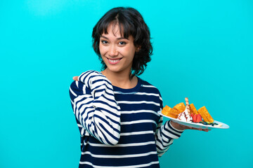 Young Argentinian woman holding waffles isolated on blue background laughing