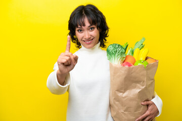 Young Argentinian woman holding a grocery shopping bag isolated on yellow background showing and lifting a finger