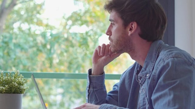 Thoughtful businessman touching chin, pondering ideas or strategy, sitting at wooden work desk with laptop, freelancer working on online project, student preparing for exam at home
