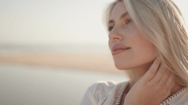 Close up of young blonde woman's face and hand as she smiles seductively at the beach with the sunset behind her.