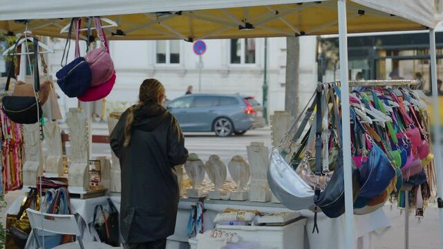 Woman vendor in street or flea market pop up shop under tent in Lisbon Portugal. Selling various beauty jewelry products and leather bags. On display in open air during spring or summer vacations fair