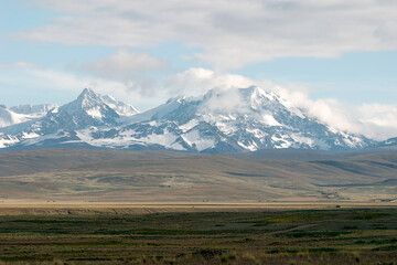 Snow capped mountains are towering over a vast plain with small villages in the distance. Clouds...