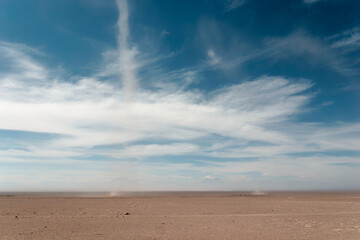 Mesmerizing dust devil forms in a deserted landscape under a stunning blue sky with fluffy clouds,...