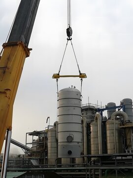 A large white tank with a conical base and flanges is lifted by a yellow crane with a long boom against a blue sky. Industrial buildings are visible in the background.