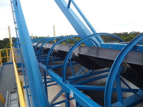 A long, black conveyor belt winds its way through a series of blue metal rollers and support structures. The conveyor belt appears to be transporting materials, likely in an industrial setting.