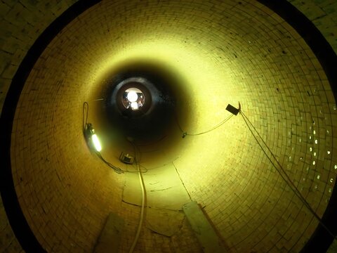 A view from inside a long, cylindrical tunnel or kiln lined with yellow bricks. The tunnel stretches into the distance, illuminated by a bright light at the far end and a few dim lights along the side